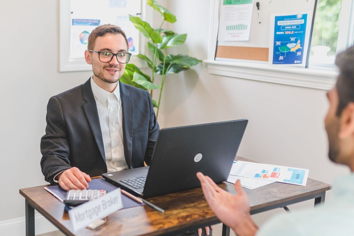 Homme en costume devant ordinateur portable, échange avec un autre homme, documents et plante sur la table