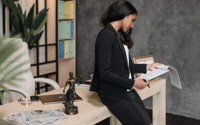 Femme en costume noir lit un document à un bureau en bois, avec une statue, un ordinateur et des papiers